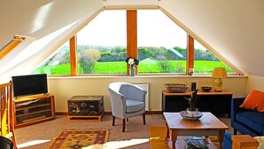 A cozy attic room with a television, a comfortable chair, a coffee table, and a view of a green landscape through large windows. - Amazing views in luxury cottage (Cheltenham)