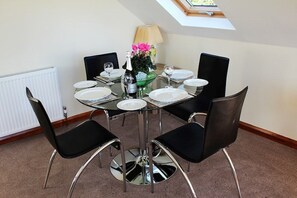 Un comedor con una mesa redonda dispuesta para cuatro personas, rodeada de sillas negras con patas cromadas. Hay un florero sobre la mesa y una lámpara se observa al fondo.