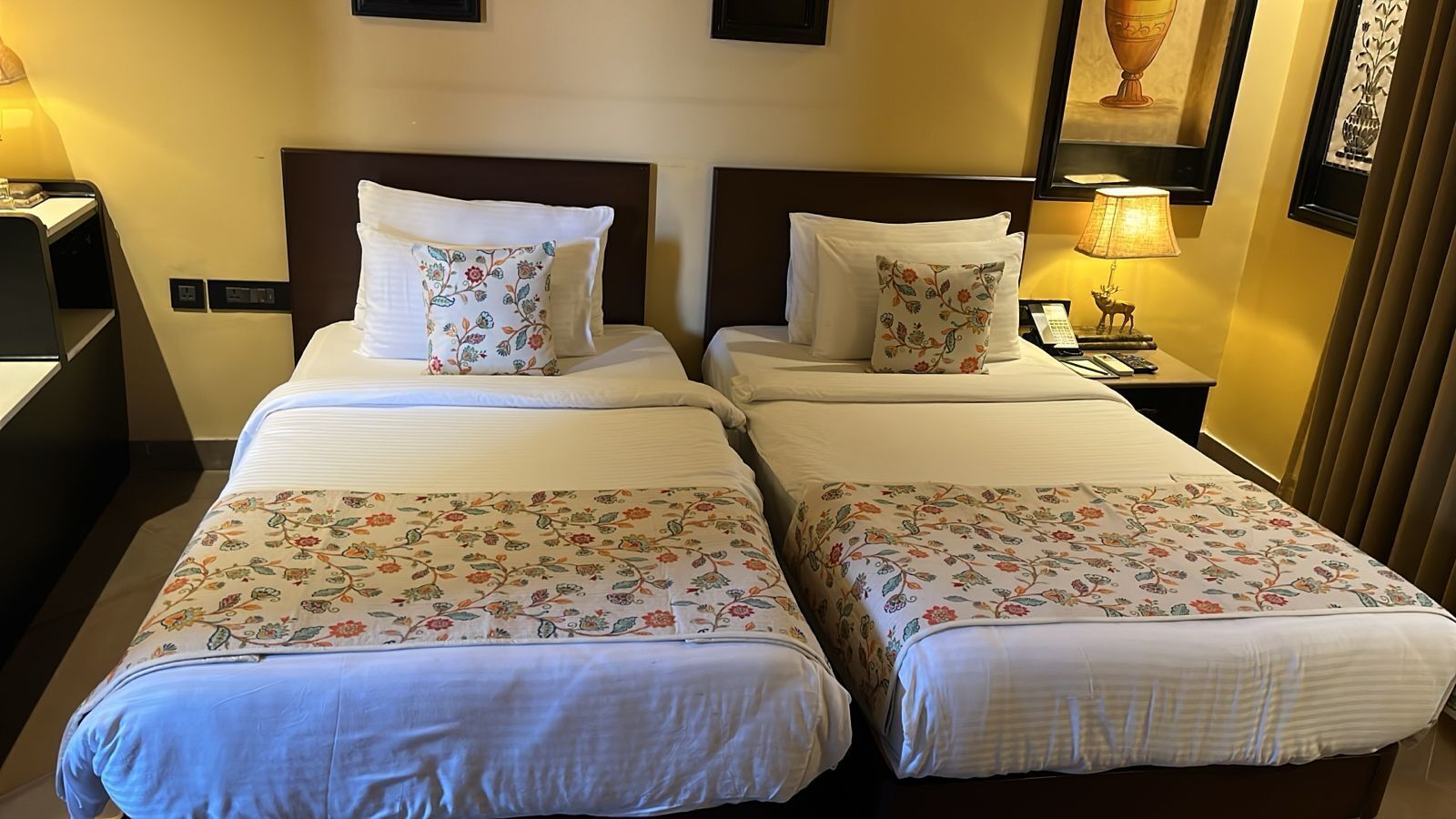 Une chambre d’hôtel avec deux lits, chacun recouvert d’une couvre-lit à motifs floraux et agrémenté de coussins assortis. On y trouve une table de chevet avec une lampe et un téléphone.