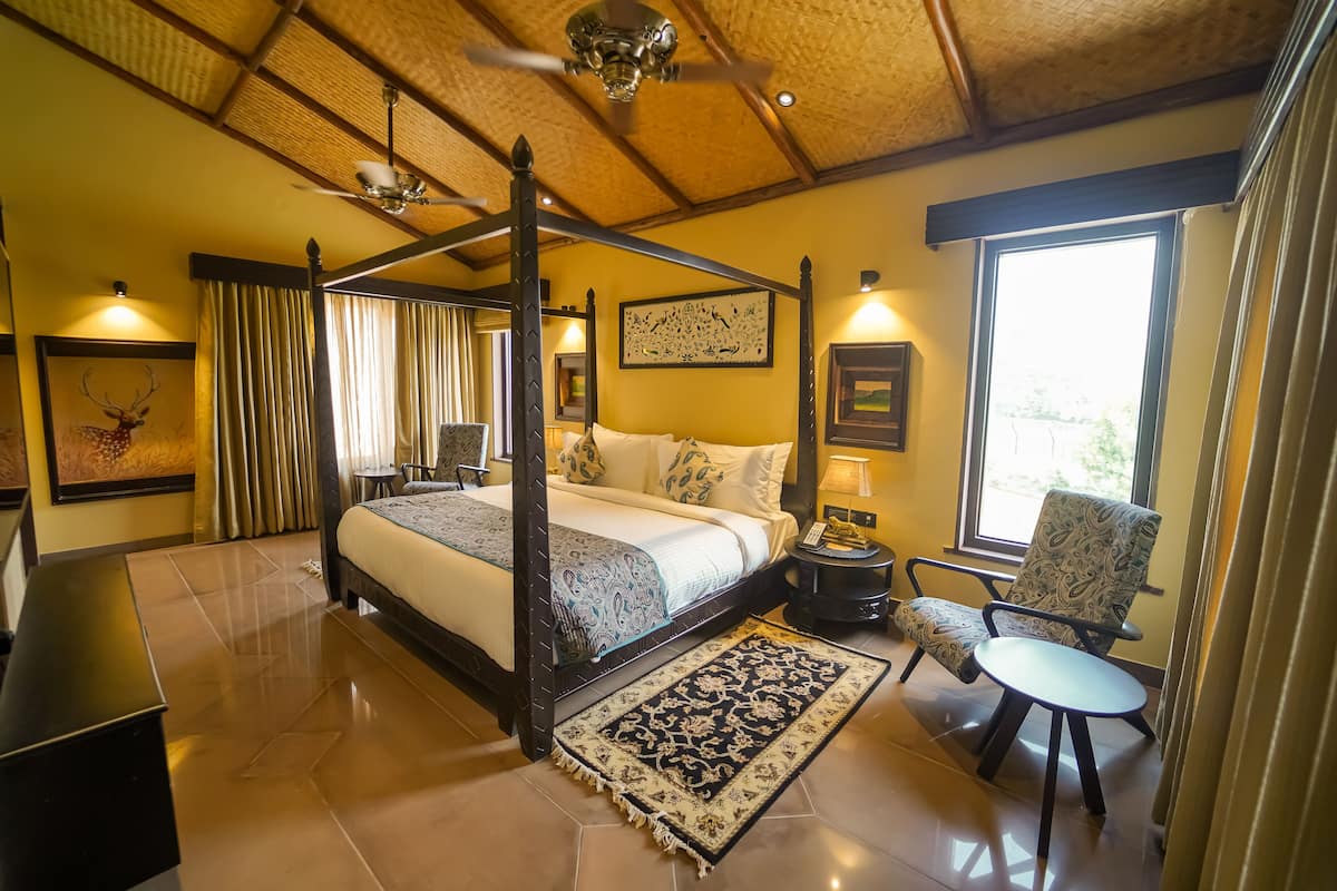 A four-poster bed with a matching canopy, a bedside table with a lamp, a chair, and a view of the outdoors through a large window.