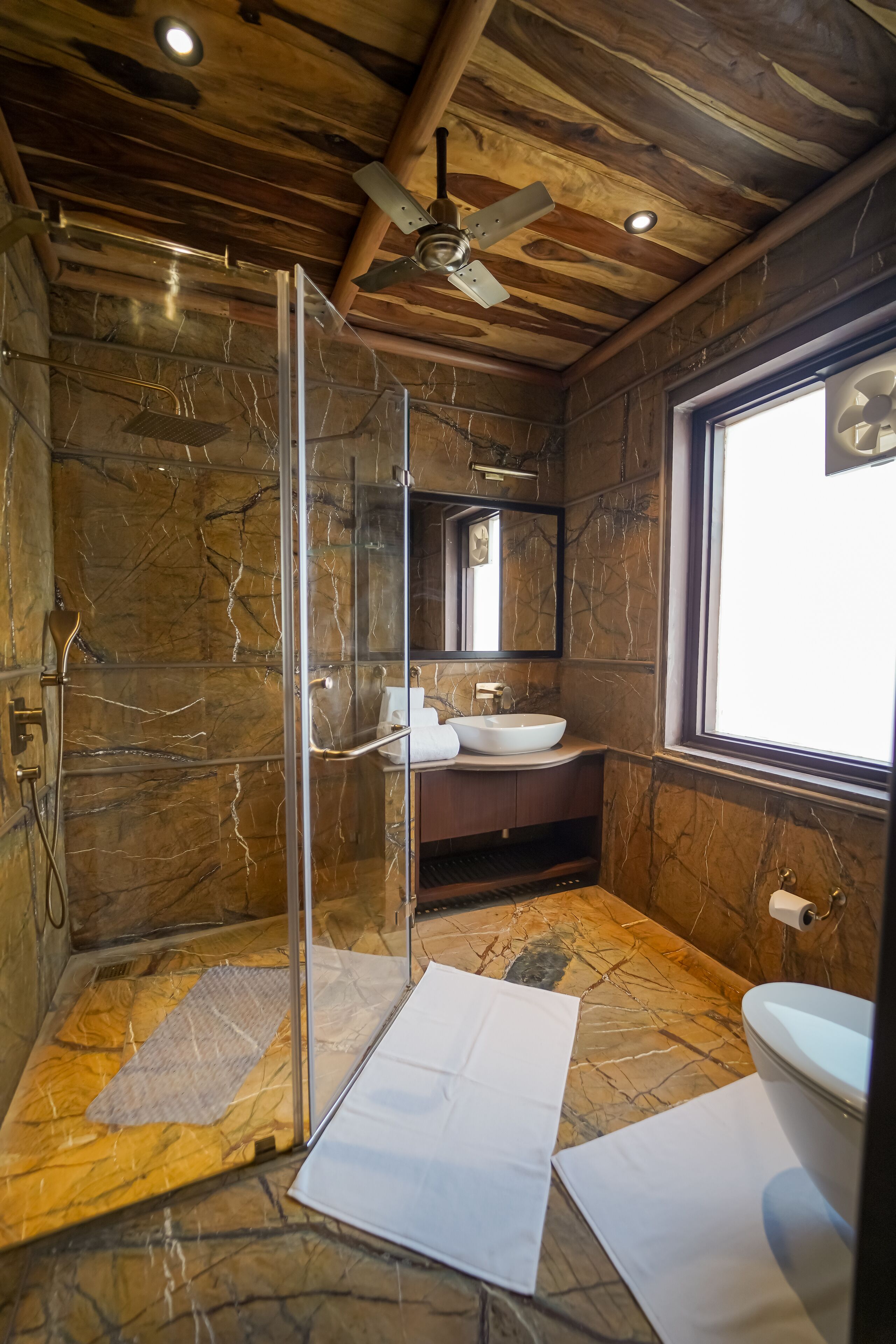 A bathroom with a glass shower enclosure, a marble-tiled floor, a white sink, and a window providing natural light.