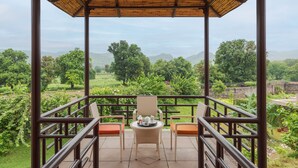 A covered outdoor seating area with a table and chairs, overlooking a lush landscape with trees and hills. - Aamaghati Wildlife Resort (Sawai Madhopur)