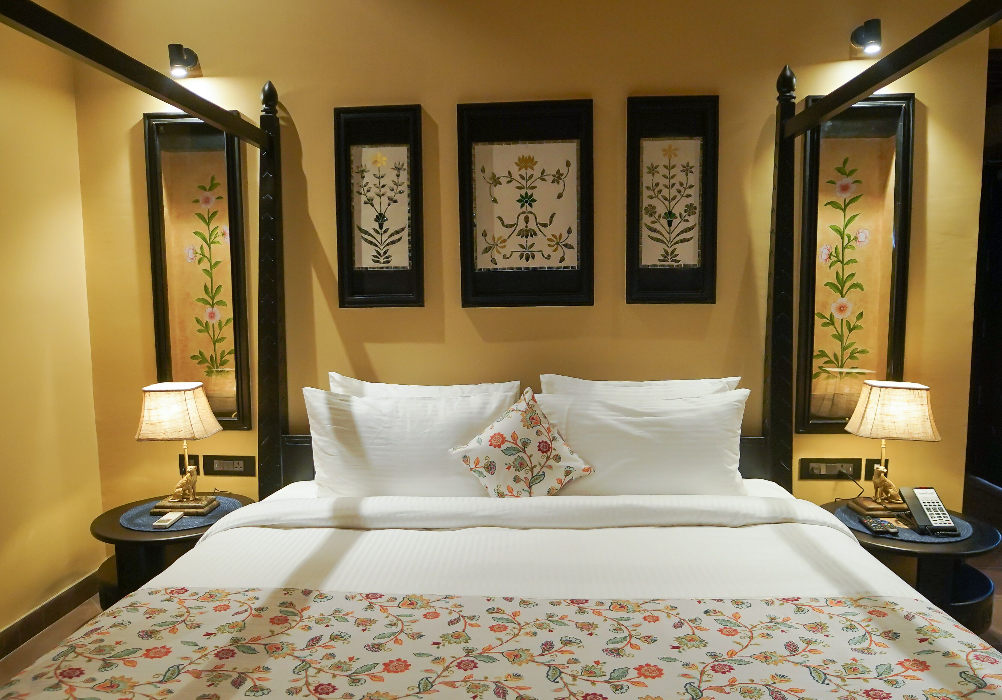 A hotel room with a large bed, white bedding, floral-patterned bedspread, two nightstands with lamps, and three framed floral artworks on the wall.