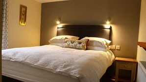 Een keurig opgemaakt bed met een witte deken en decoratieve kussens, een nachtkastje met een lamp en een ingelijste foto aan de muur.
