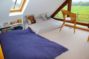 Een slaapkamer met een schuine kap, een bed met een paarse deken, een houten stoel, een boekenplank en een raam met uitzicht op een groen veld.