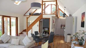 Een ruime woonkamer met een bank, een eettafel en stoelen, een trap naar een hoger gelegen verdieping, en een keukenhoek.