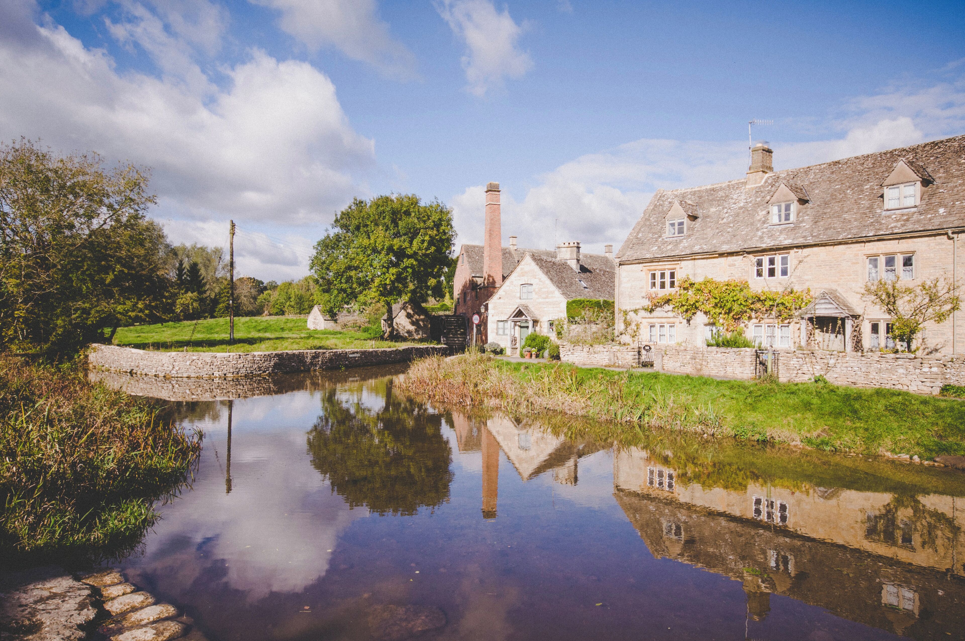 A traditional stone building with a reflection in a calm water body, surrounded by greenery.
