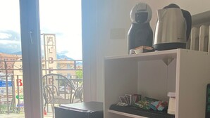 Una habitación con un mueble blanco con estantes que contiene una cafetera, un hervidor y algunos alimentos envasados. Hay un cubo de basura pequeño y un microondas en el suelo.