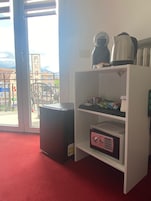 A room with a white shelving unit containing a coffee maker, a kettle, and some packaged food. There is a small trash bin and a microwave on the floor.