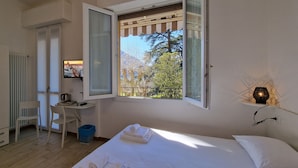 Un dormitorio con cama, escritorio y silla, un radiador, y ventana con vistas a árboles y un edificio.