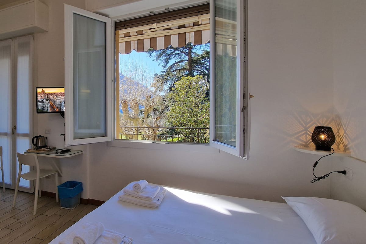 A bedroom with a bed, a desk with a chair, a radiator, and a window with a view of trees and a building.