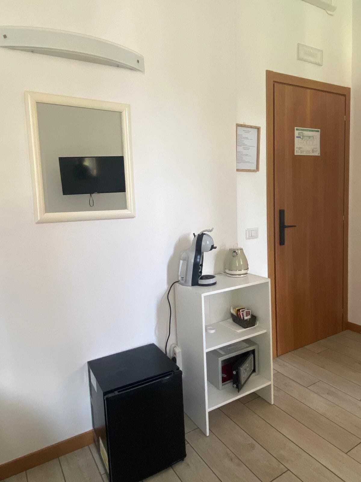 A room with a television mounted on the wall, a coffee machine on a white shelf, a black refrigerator, and a wooden door with a sign.