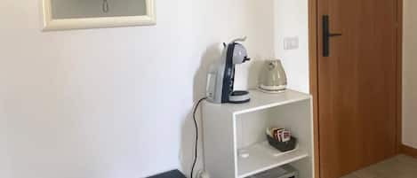 Une pièce avec un téléviseur fixé au mur, une machine à café posée sur une étagère blanche, un réfrigérateur noir et une porte en bois avec une pancarte.