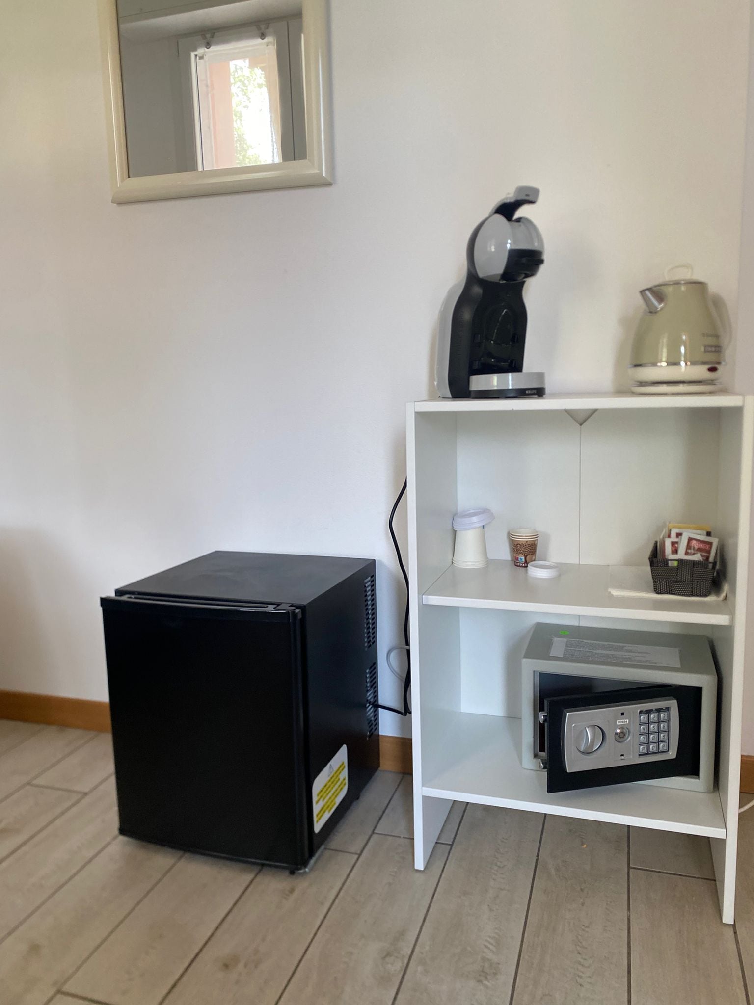 A room with a white shelf holding a coffee maker, a kettle, and a safe. There is a black refrigerator on the floor.