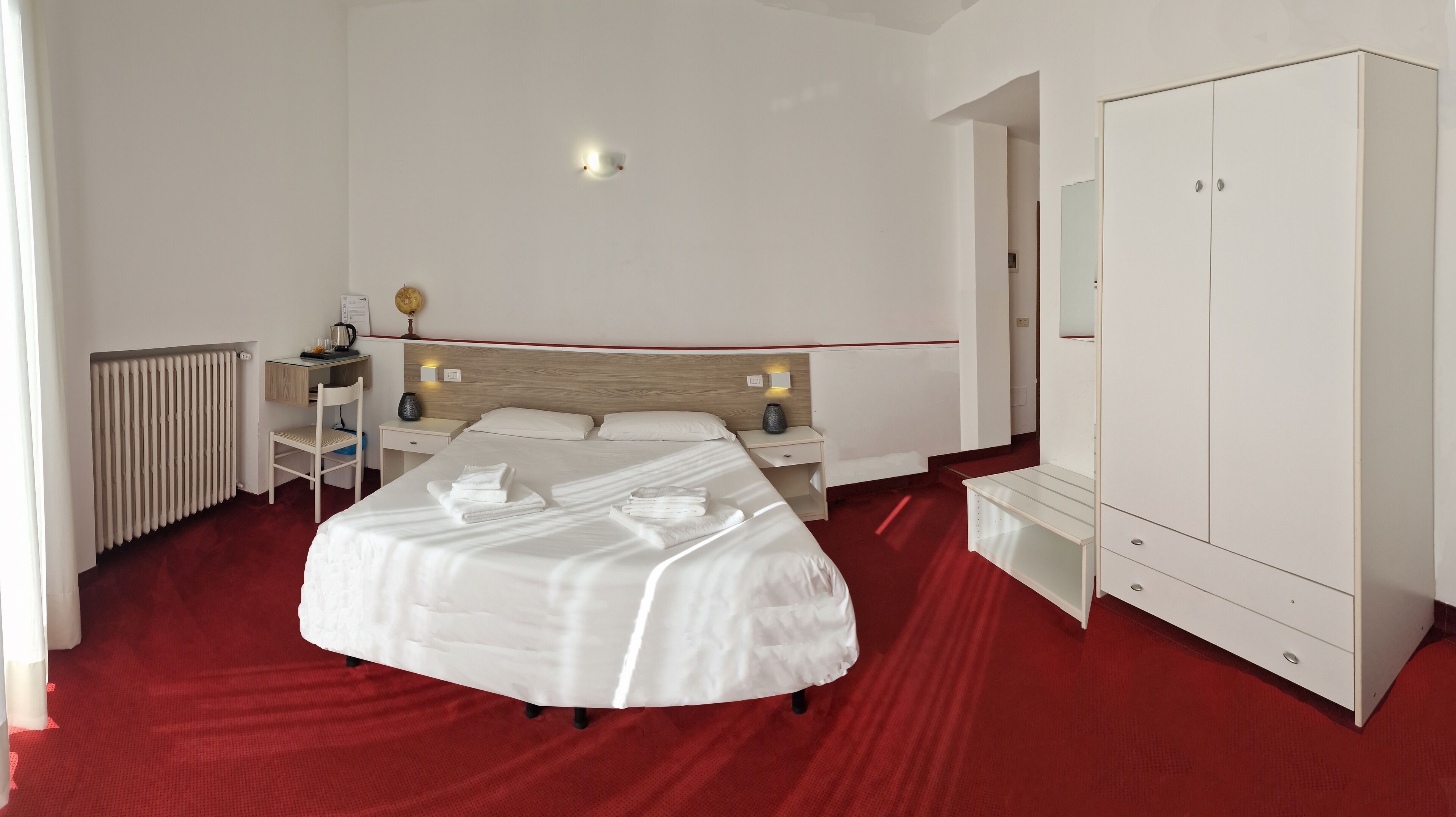 A hotel room with a large bed, a desk with a chair, a wardrobe, and a radiator.