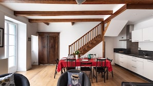 Un comedor con mantel rojo, vigas de madera, una escalera y una cocina con armarios blancos.