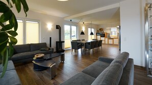 Amplia sala de estar con sofá, mesa de comedor y sillas. Se observa una chimenea y una cocina al fondo.