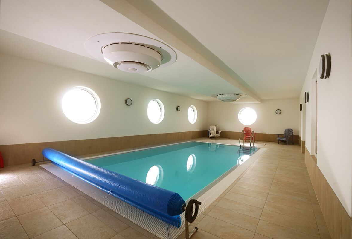 A large indoor swimming pool with a blue pool ladder, circular windows, and ceiling vents.