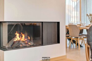 Un salon moderne doté d’une cheminée intégrée, un coin repas avec des meubles en bois et une vue sur la ville grâce à de grandes fenêtres.