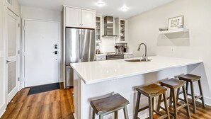 A modern kitchen with white cabinetry, a stainless steel refrigerator, a built-in oven, and a large island with bar stools.