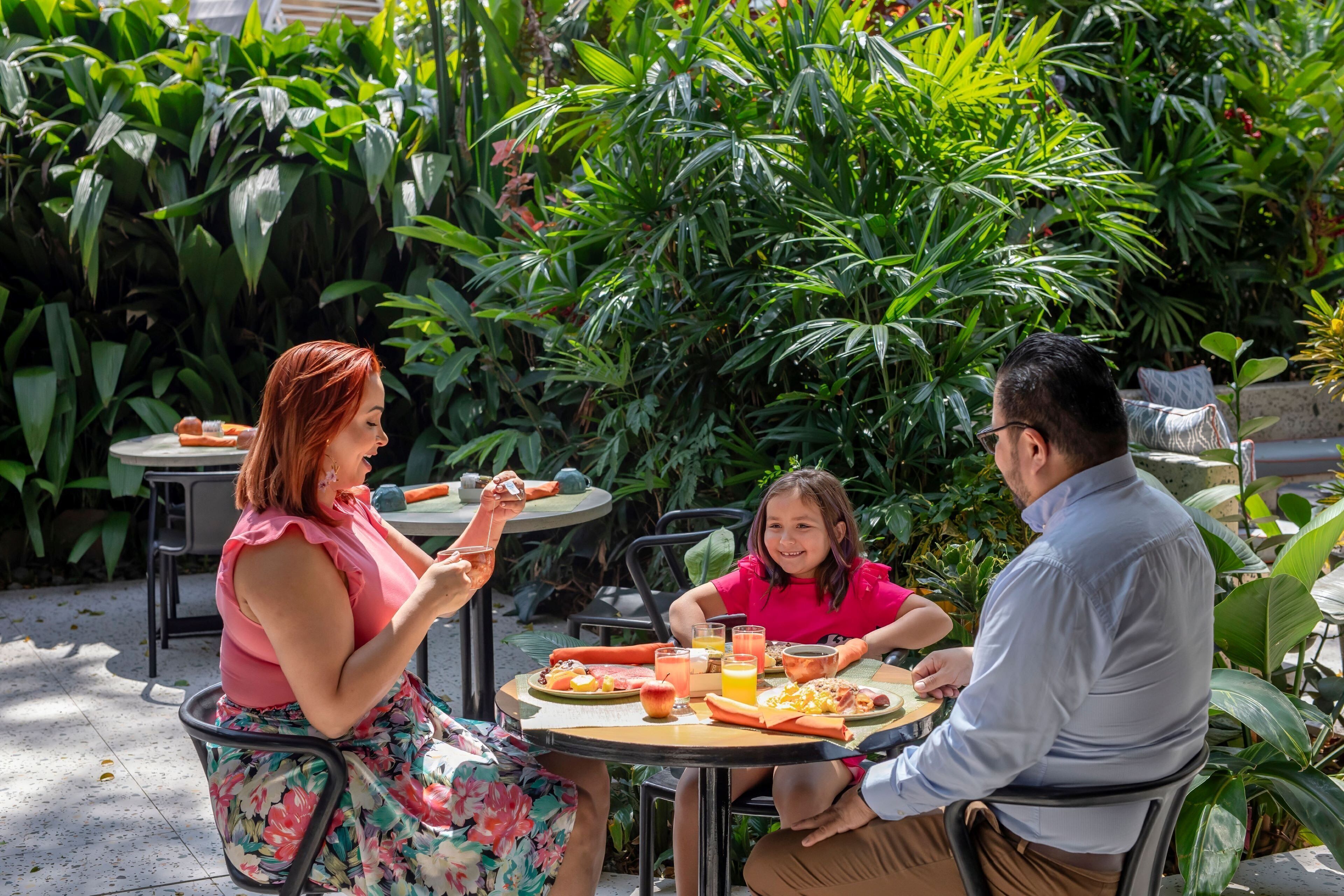 A family dining outdoors at a table surrounded by lush greenery.