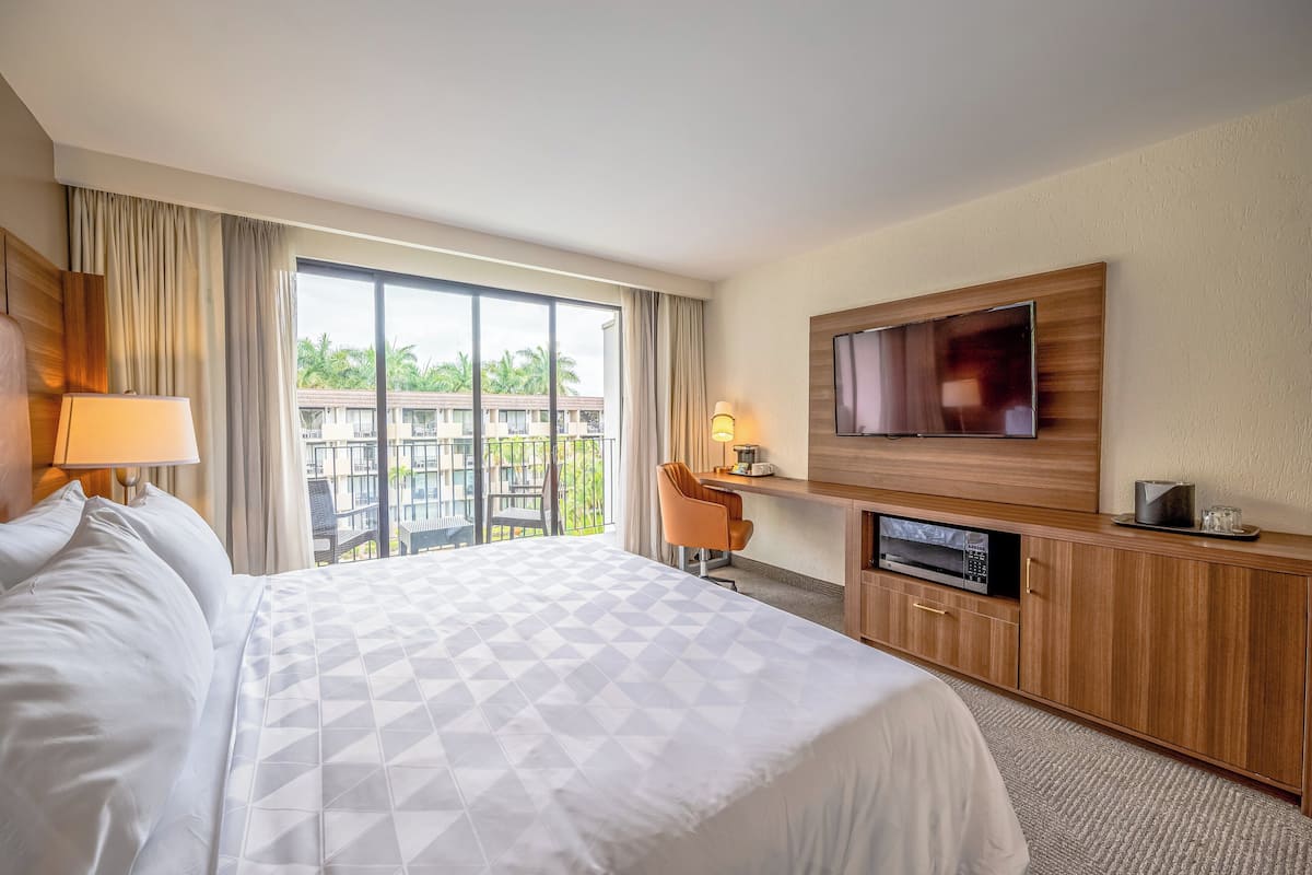 Ein Hotelzimmer mit einem großen Bett, einem Schreibtisch mit Stuhl, einem Flachbildfernseher und Blick auf einen Balkon mit Palmen.