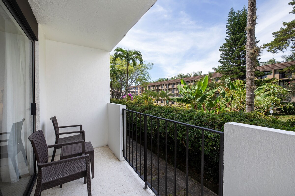 A balcony with wicker furniture, a view of a landscaped garden, and a building in the background.