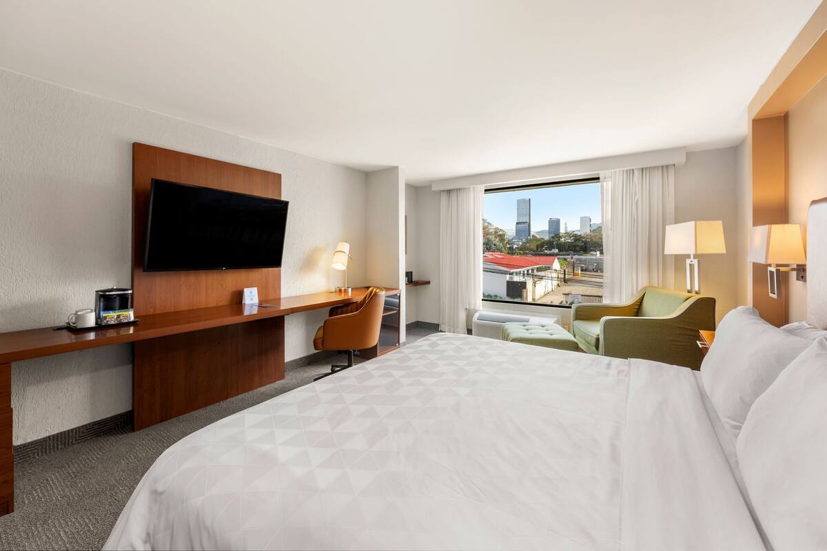 Ein Hotelzimmer mit einem großen Bett, einem Schreibtisch mit Flachbildfernseher, einem Sessel, einem kleinen Tisch und Blick auf die Stadtlandschaft durch das Fenster.