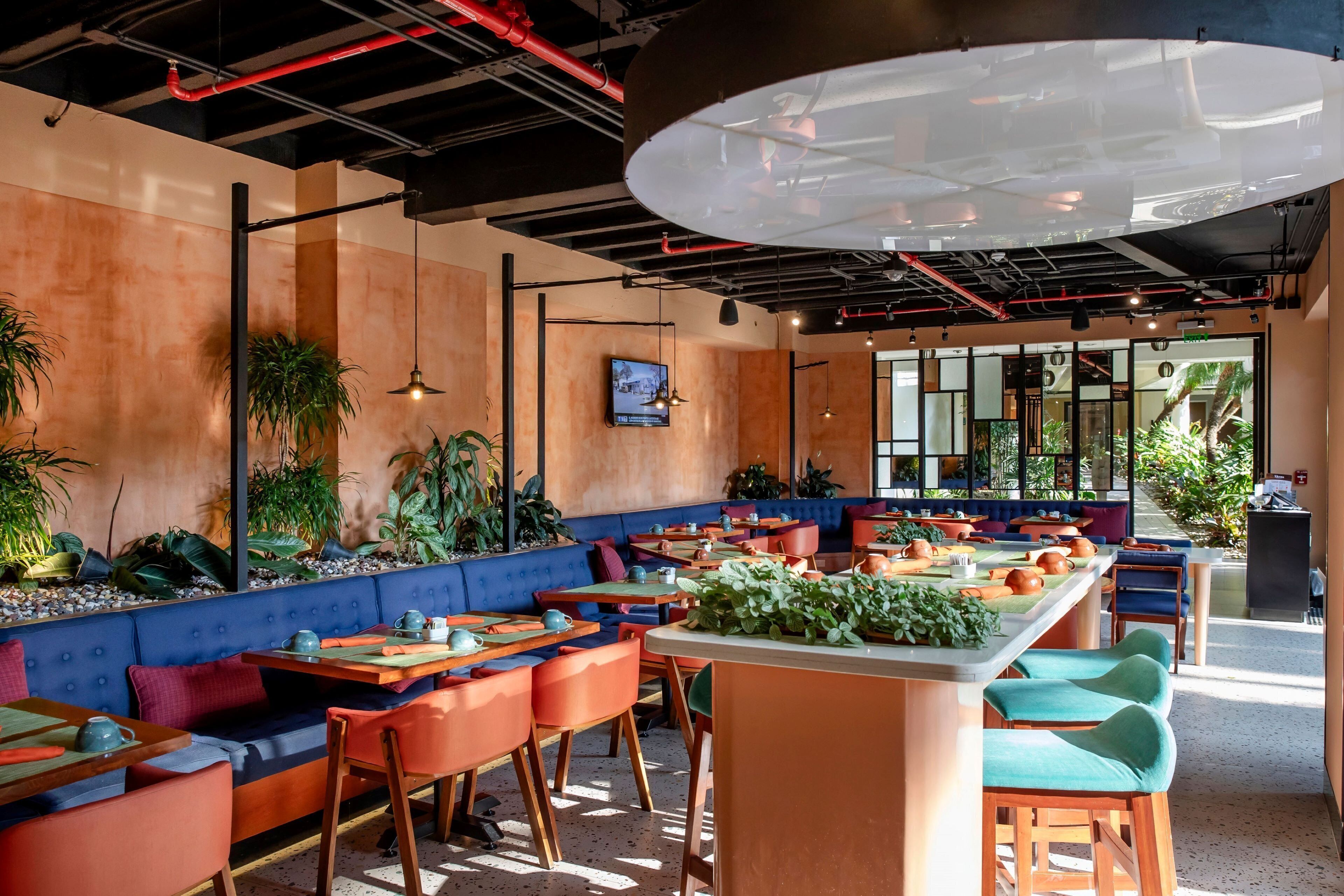 Un restaurante moderno con asientos azules y anaranjados, una isla central grande con plantas y un televisor en la pared.