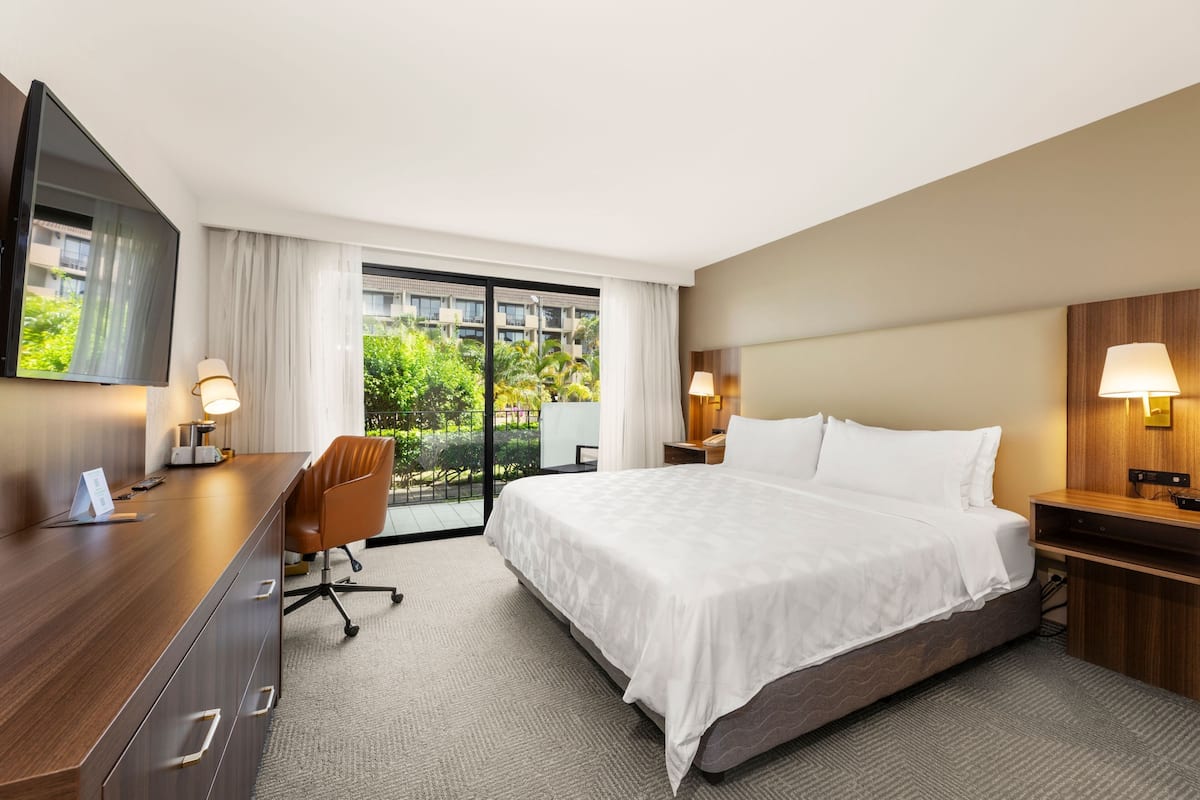 Ein Hotelzimmer mit einem großen Bett, einem Schreibtisch mit Stuhl, einem Fernseher und einem Balkon mit Blick ins Grüne.