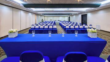 A conference room with rows of blue chairs, a long table with a blue tablecloth, and white flower arrangements on the table.