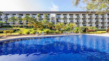A swimming pool with lounge chairs, surrounded by palm trees and a multi-story building with balconies.