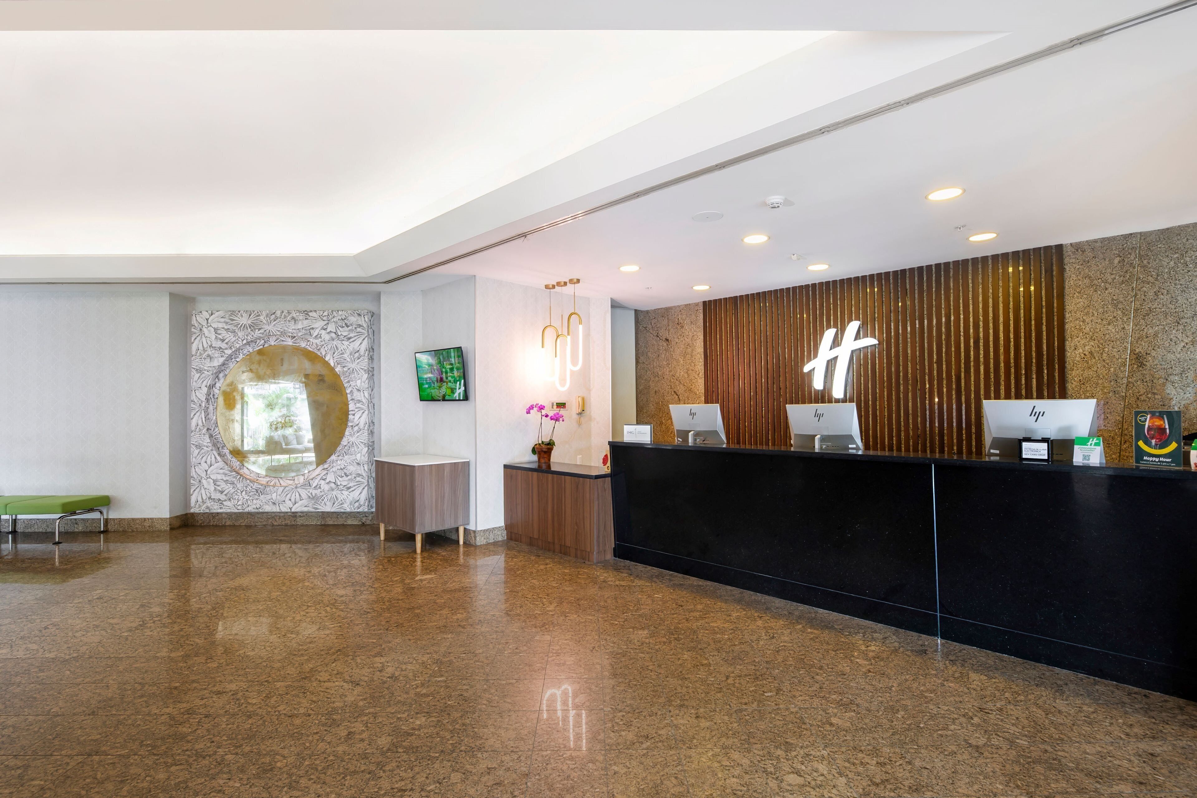 A hotel reception area with a modern design, featuring a black reception desk, a large circular mirror, and a wooden accent wall.