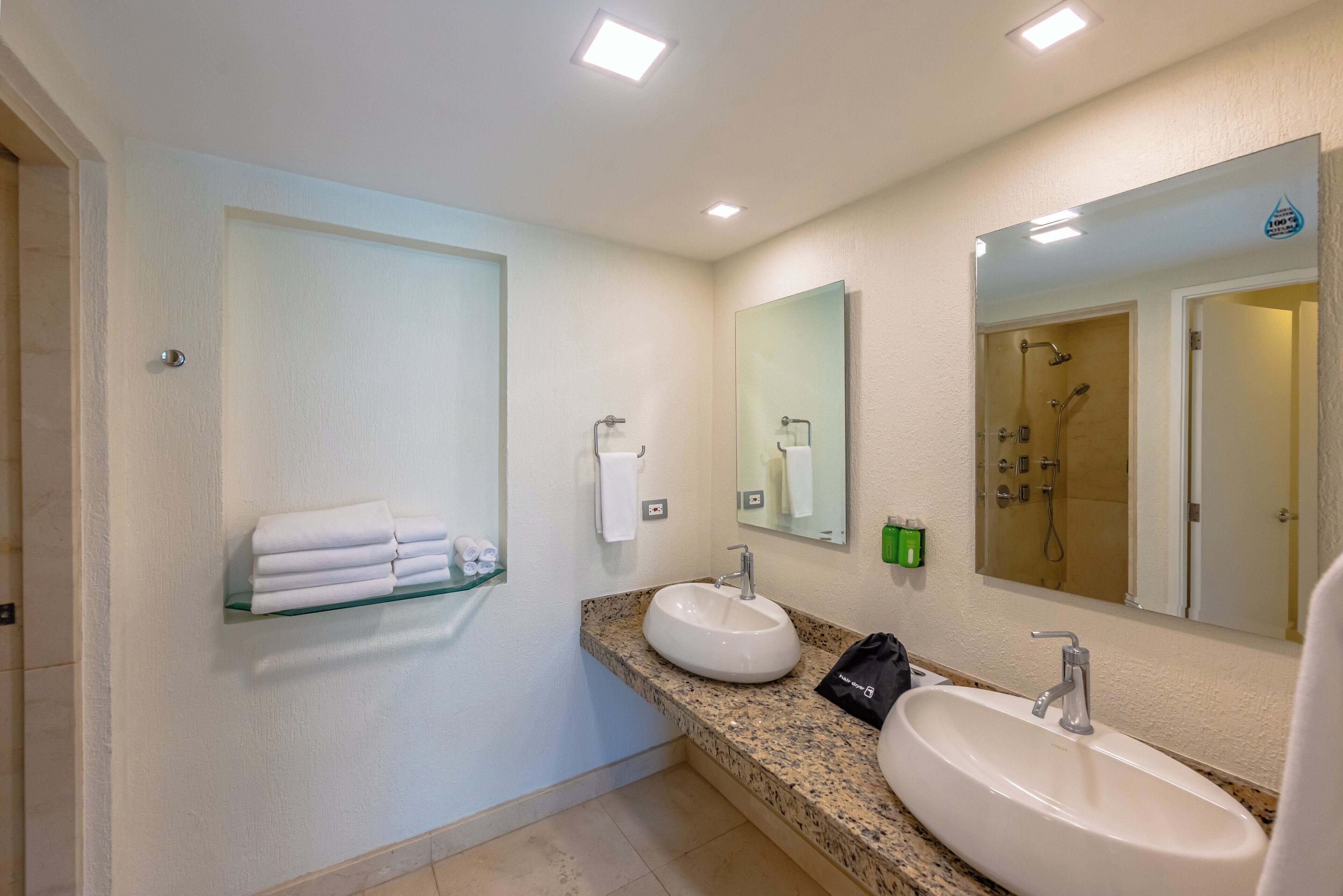 A bathroom with two sinks, a glass shelf with folded towels, a mirror, and a shower area.