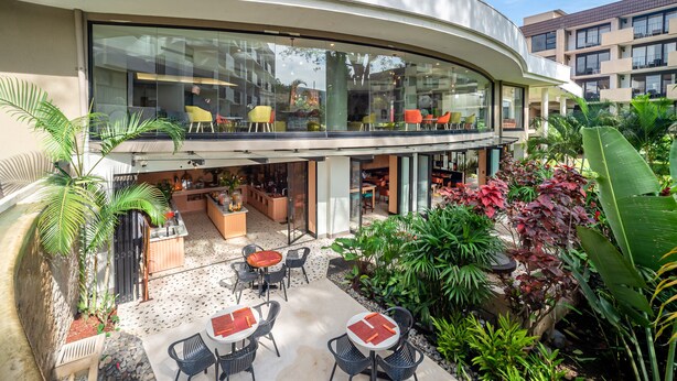 A modern hotel with a curved glass facade, outdoor seating area with tables and chairs, and lush greenery. - Holiday Inn San Jose La Sabana by IHG (San José)