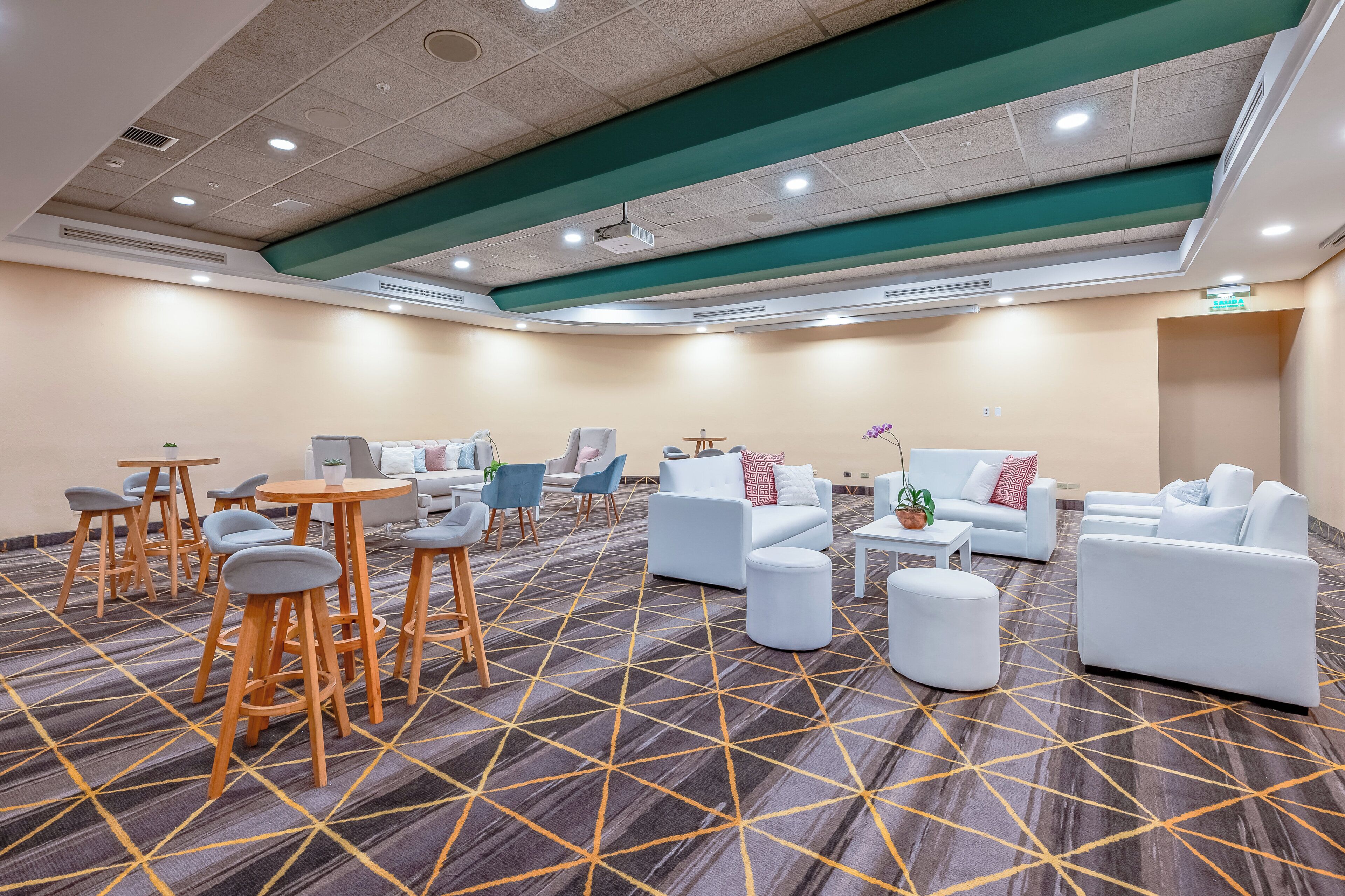 A hotel lobby with a carpeted floor, seating areas with tables and chairs, and a reception desk.
