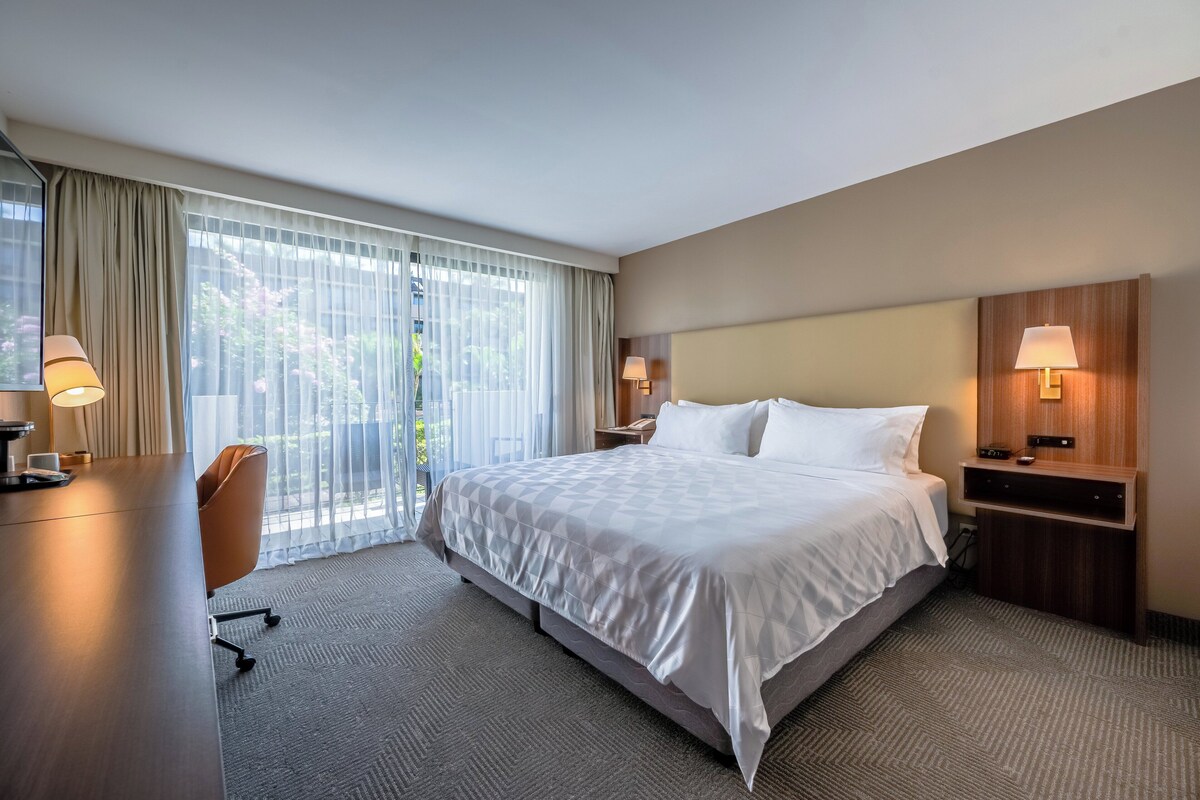 A hotel room with a large bed, a desk, a chair, two bedside tables with lamps, and a view of greenery through the window.