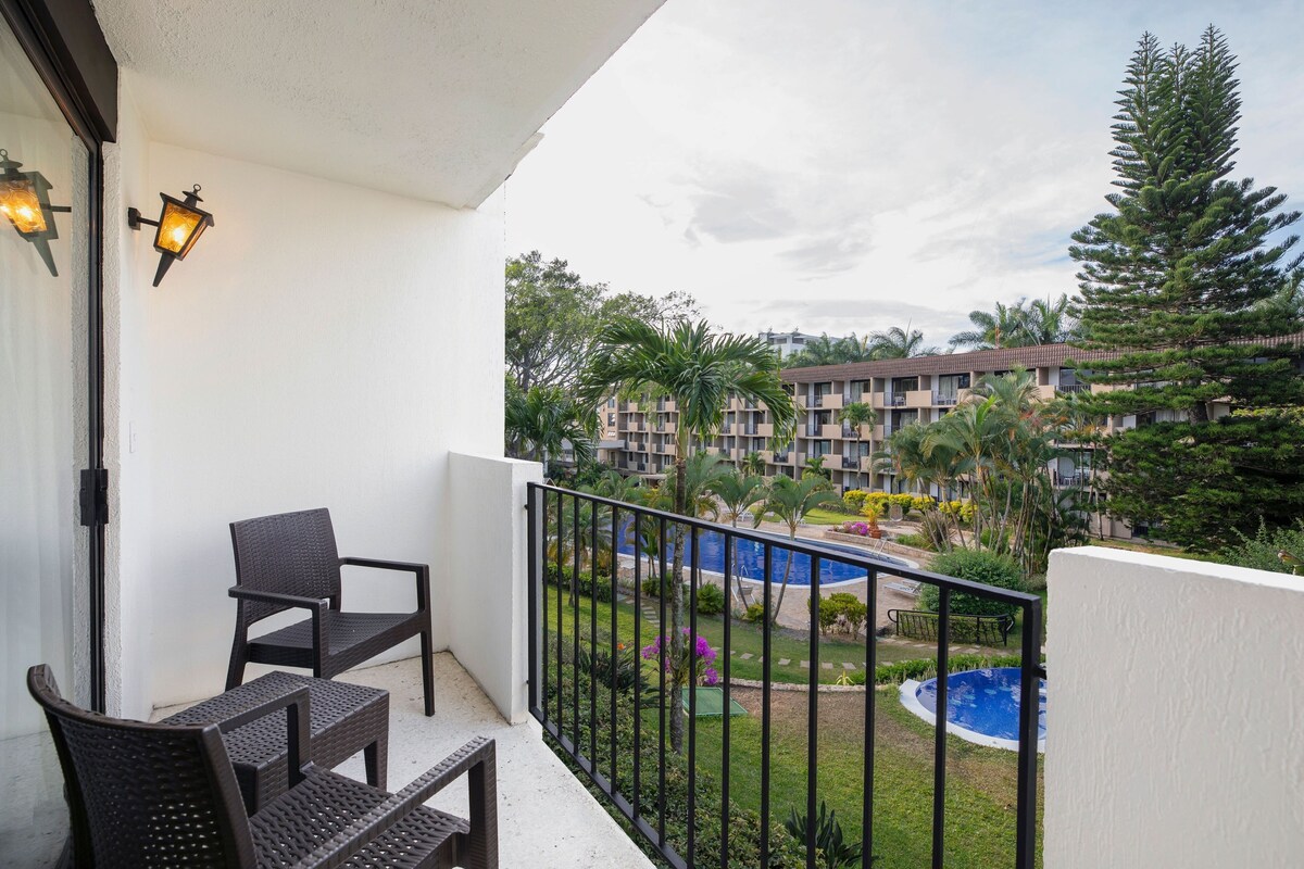 A balcony with wicker chairs, a pool, and a view of a multi-story building with a garden.