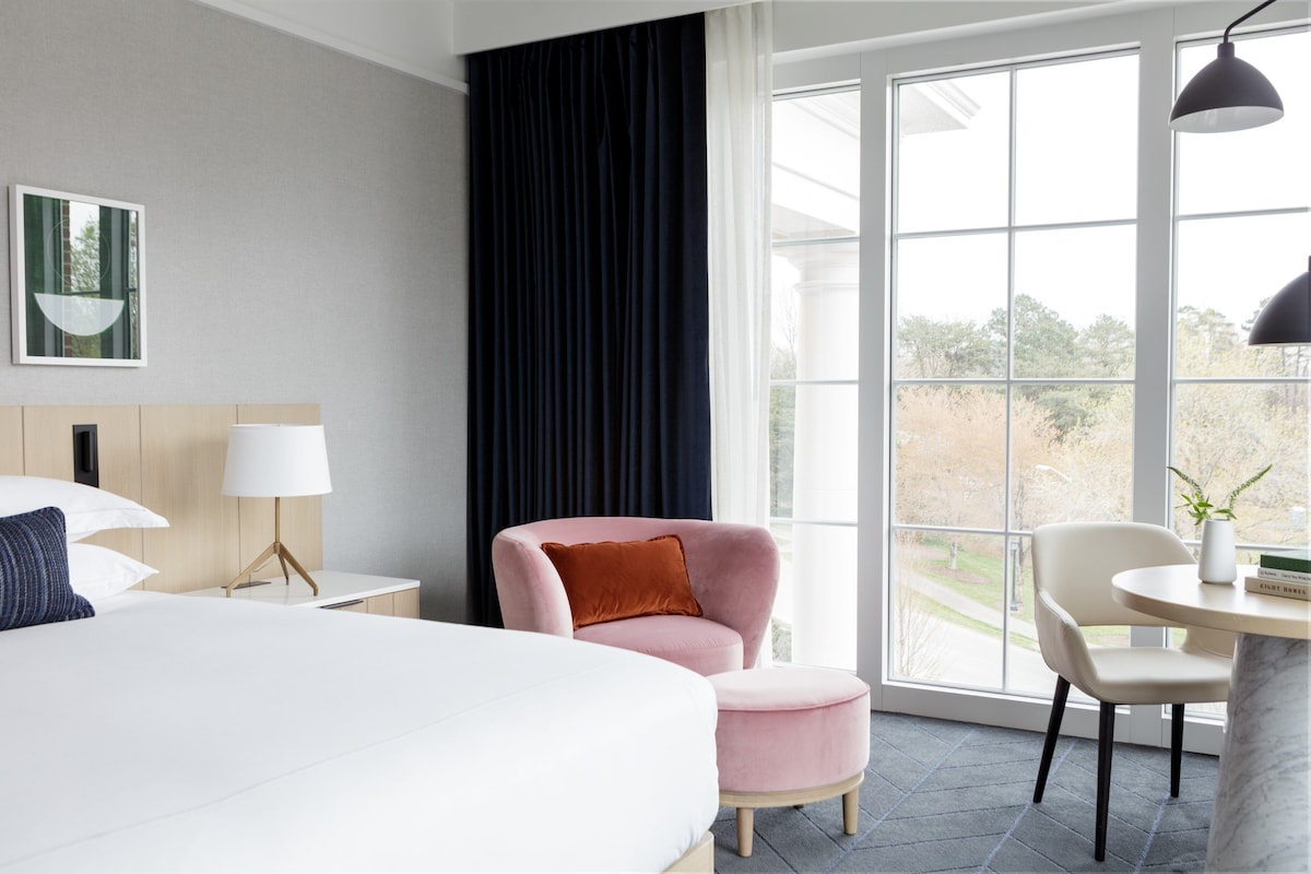 A hotel room with a bed, a chair, a small table, and a window with a view.