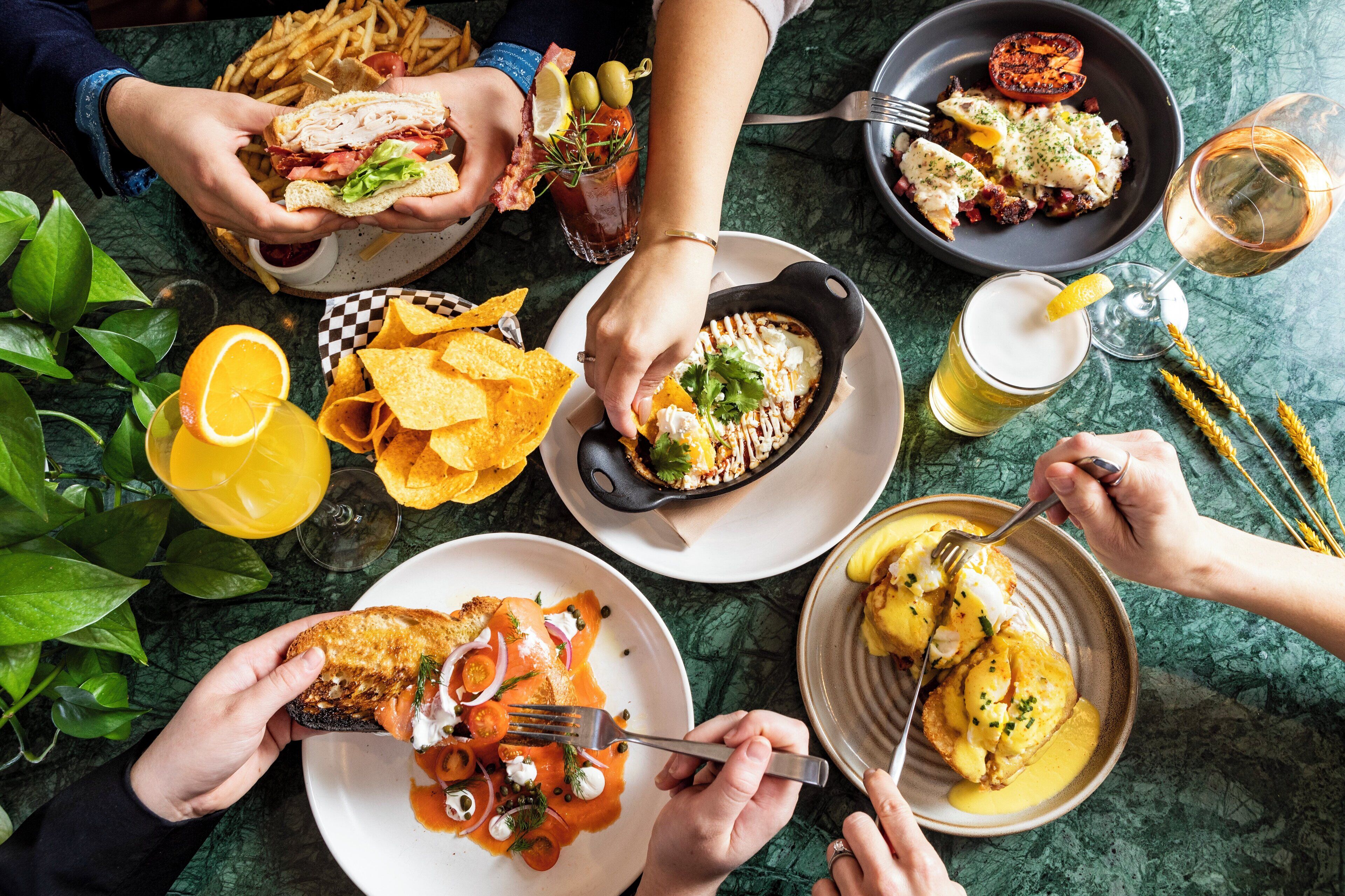 Una mesa de comedor con varios platos, incluyendo un sándwich, papas fritas y un guiso en sartén, además de bebidas y una planta.