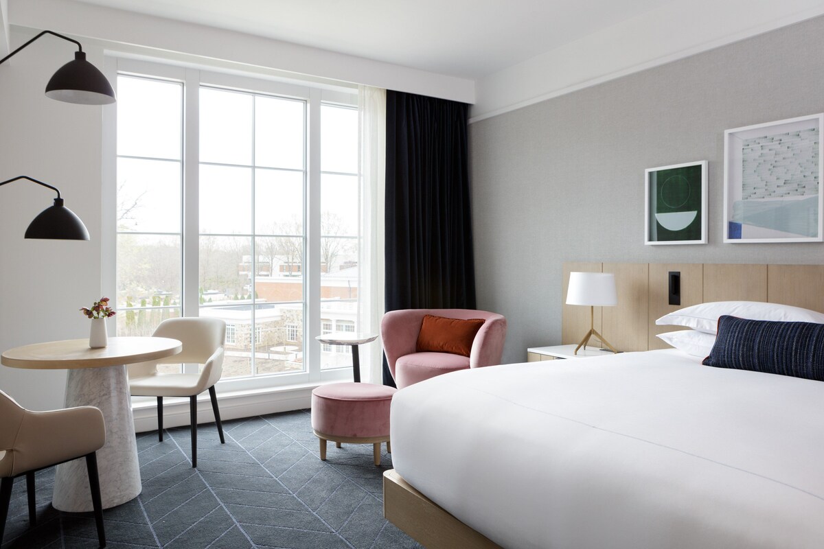 A hotel room with a large bed, a desk, a chair, a small table, and a window with a view of buildings.