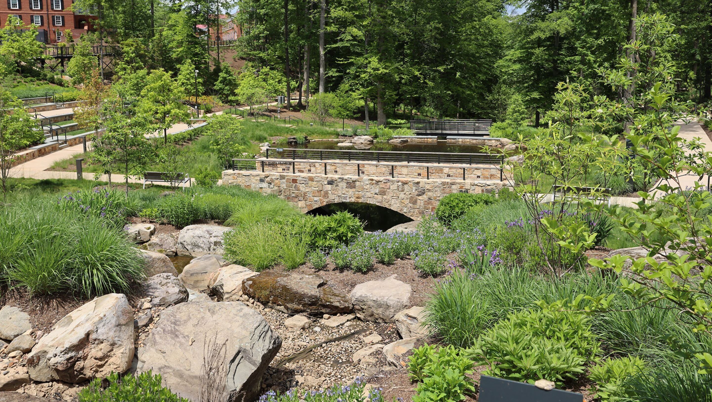 A stone bridge over a stream in a landscaped garden with various plants and a stone pathway.