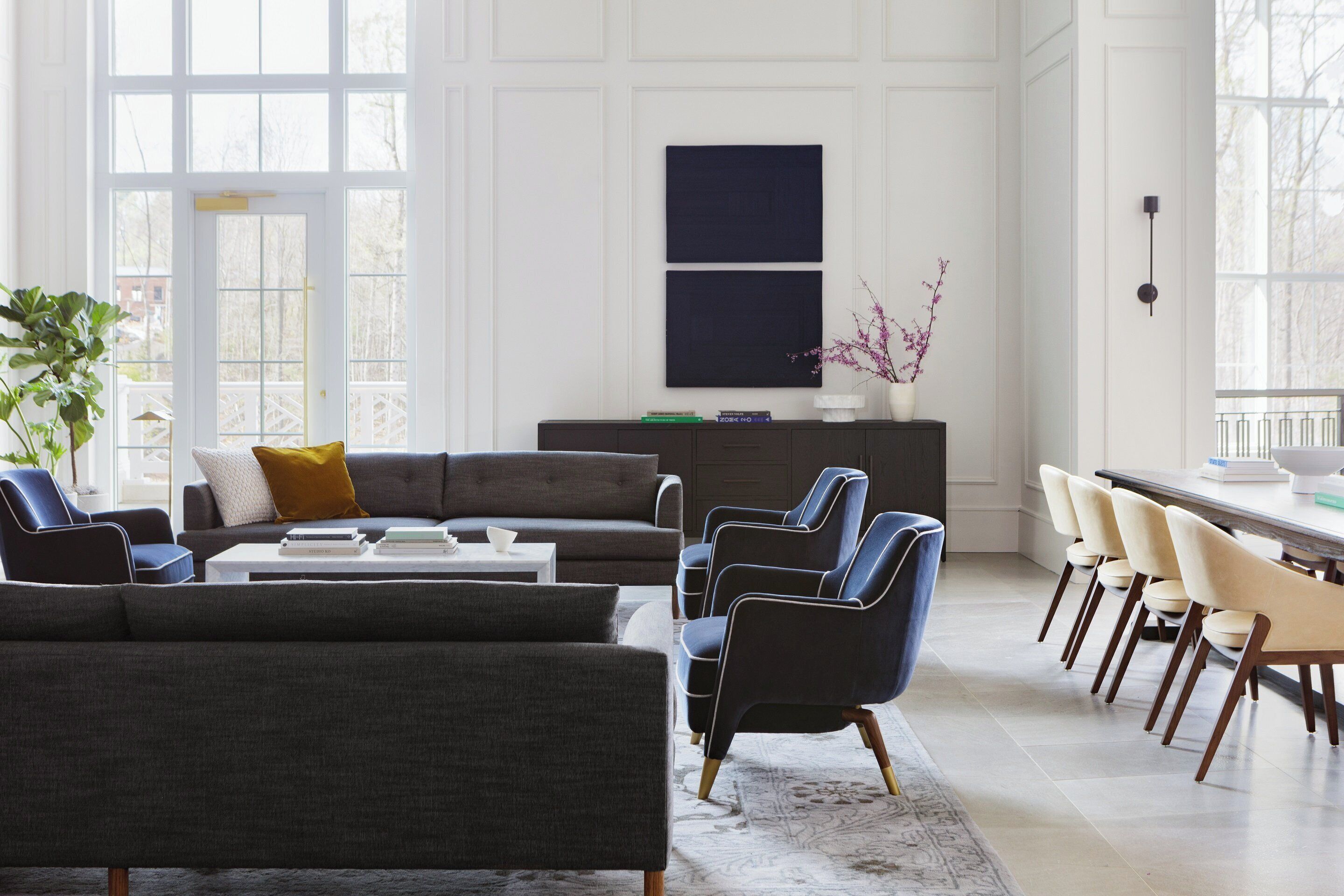 A modern living room with a gray sectional sofa, a coffee table, and a dining area with chairs. There is a white cabinet with books and a vase with flowers.