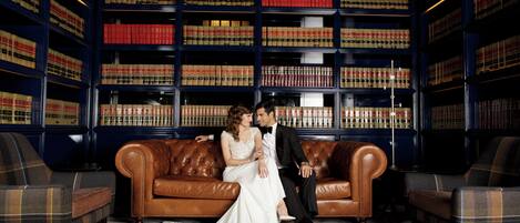 A couple on a brown leather couch in a library with bookshelves in the background.