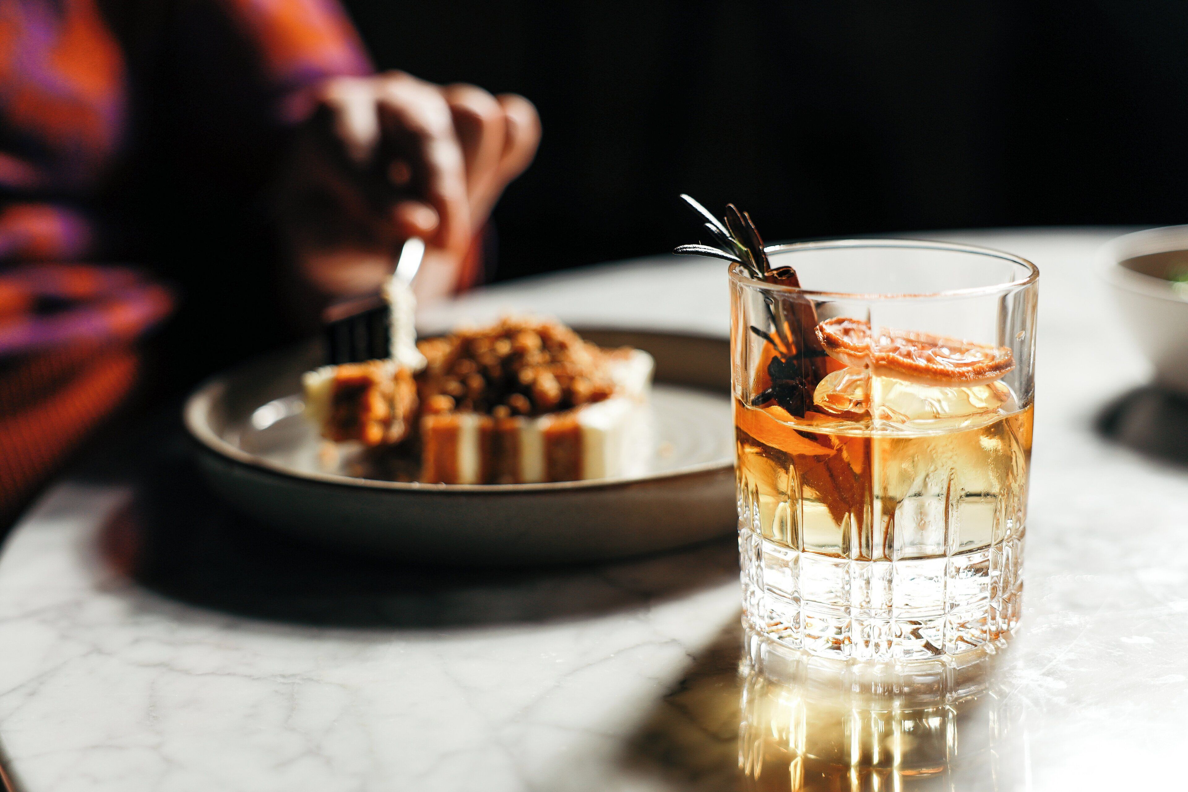 A glass of a clear beverage with a slice of orange and a sprig of rosemary, accompanied by a plate of food in the background.