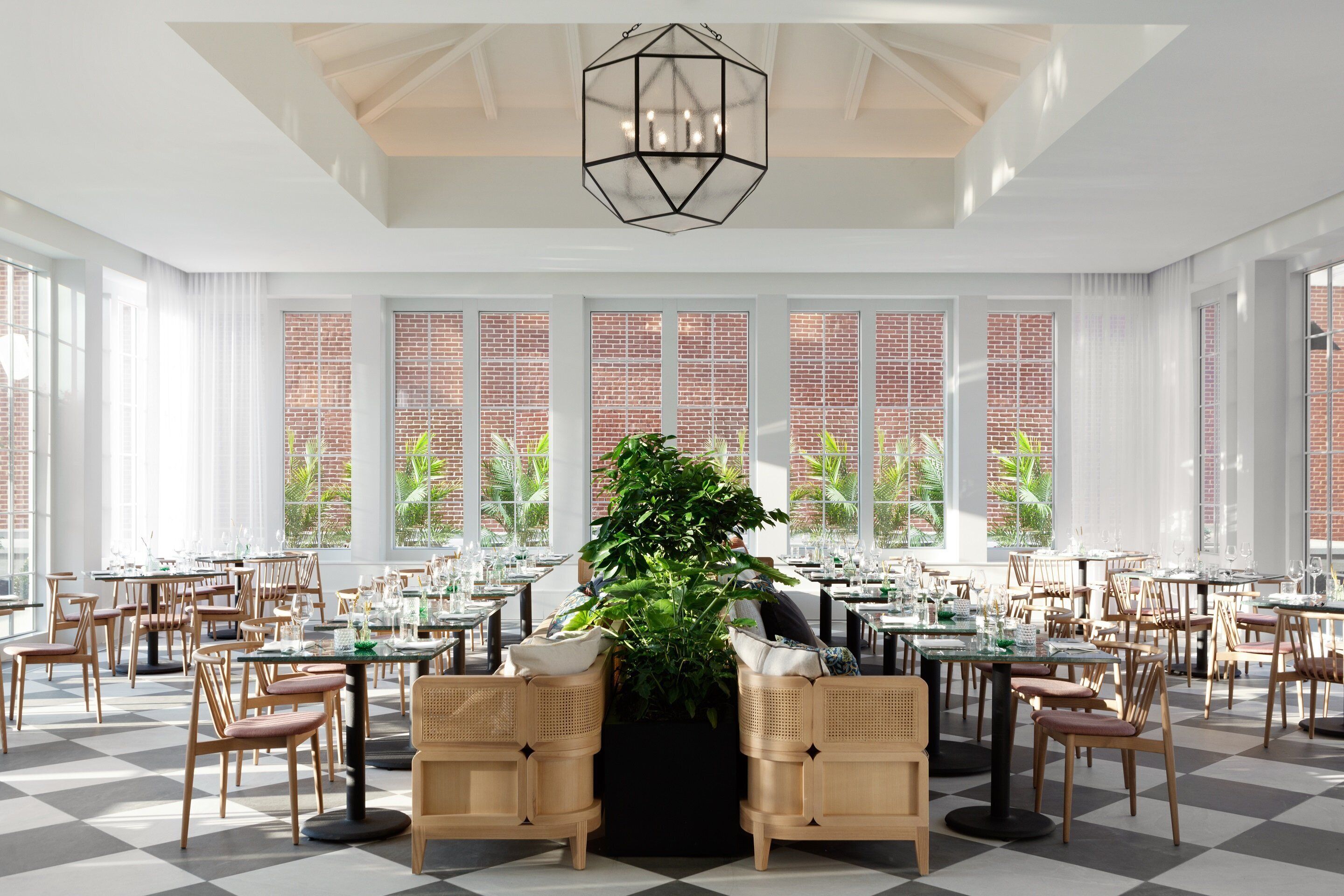 A spacious dining area with high ceilings, large windows, and a central plant. Tables and chairs are arranged for dining.