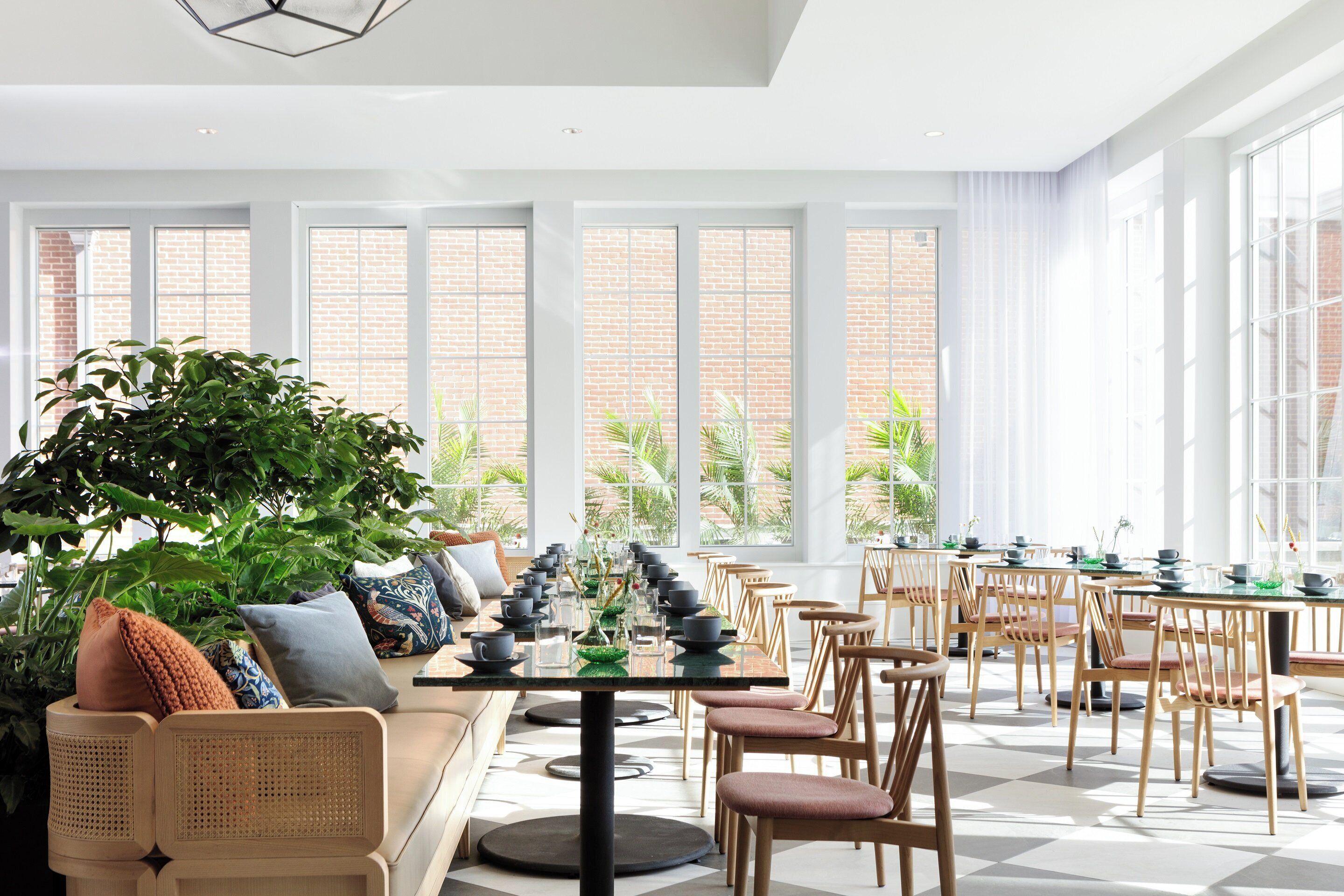 A spacious indoor area with a long table set for dining, surrounded by wooden chairs, and large windows offering views of a brick building and greenery outside.