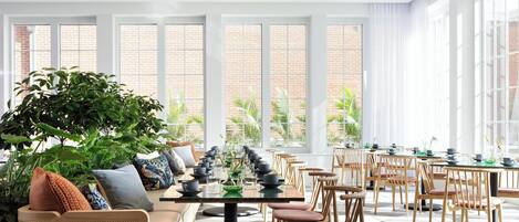 A spacious indoor area with a long table set for dining, surrounded by wooden chairs, and large windows offering views of a brick building and greenery outside.