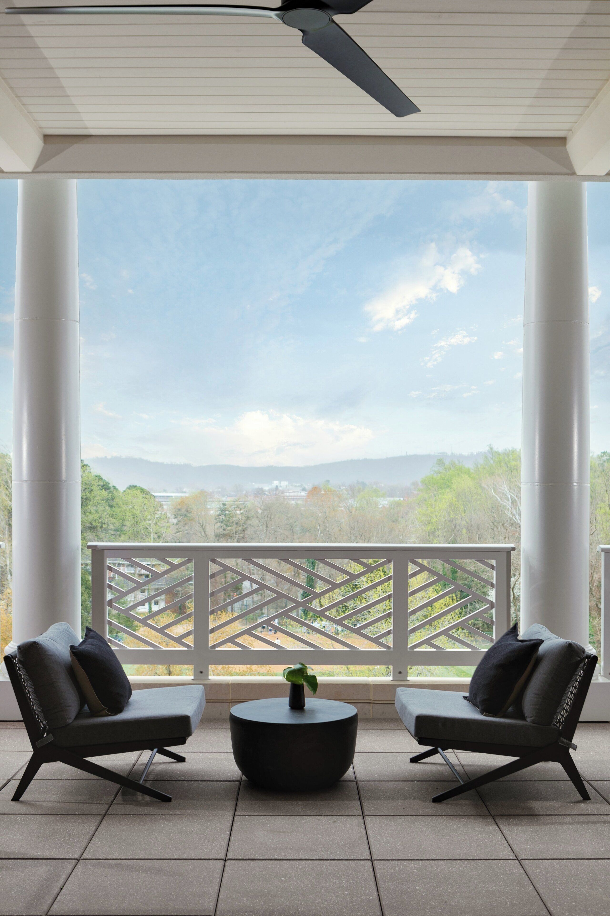 A covered outdoor seating area with two chairs and a round table, overlooking a scenic landscape with trees and distant hills.