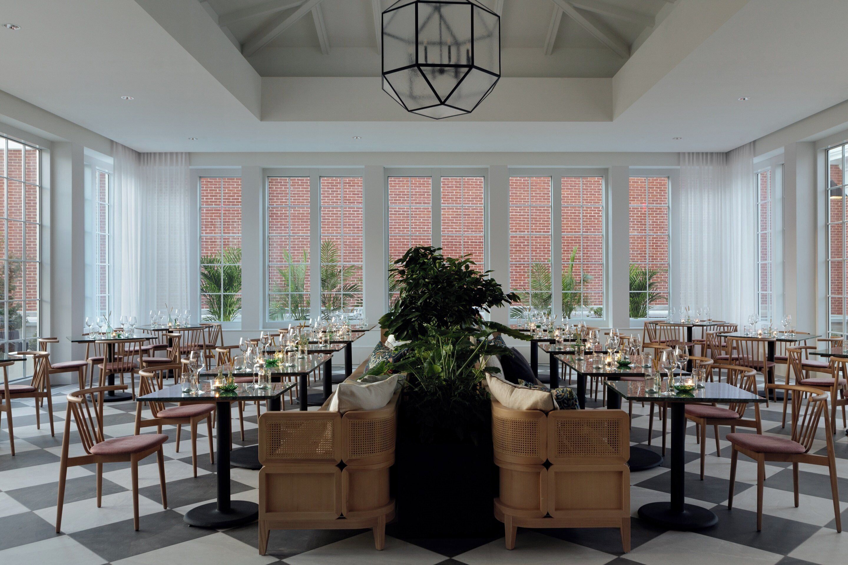 A spacious dining area with high ceilings, large windows, and a central plant. Tables are set with chairs, and the floor has a checkered pattern.